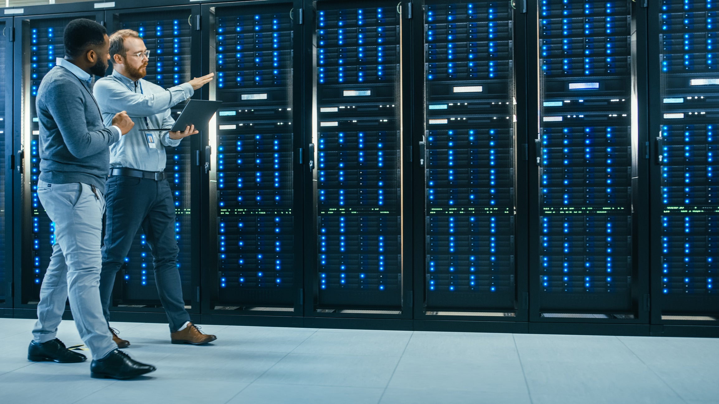 it-technician-with-a-laptop-computer-and-engineer-colleague-are-talking-in-data-center-while-walking-next-to-server-racks-running-diagnostics-or-doing-maintenance-work